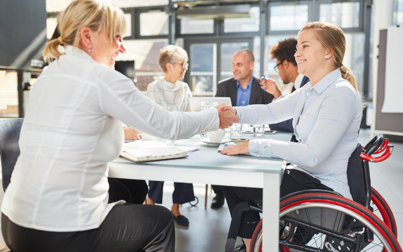 Two women shaking hands during a meeting. One young white woman is in a electric wheelchair at a table while preparing for the meeting. A full team surrounds her and shares her enthusiasm for the work they're doing.